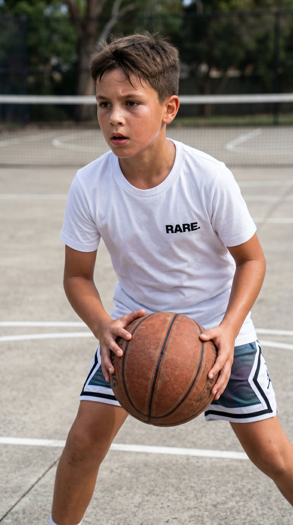 Child holding a basketball on a court wearing a white t-shirt with 'RARE' printed on it.