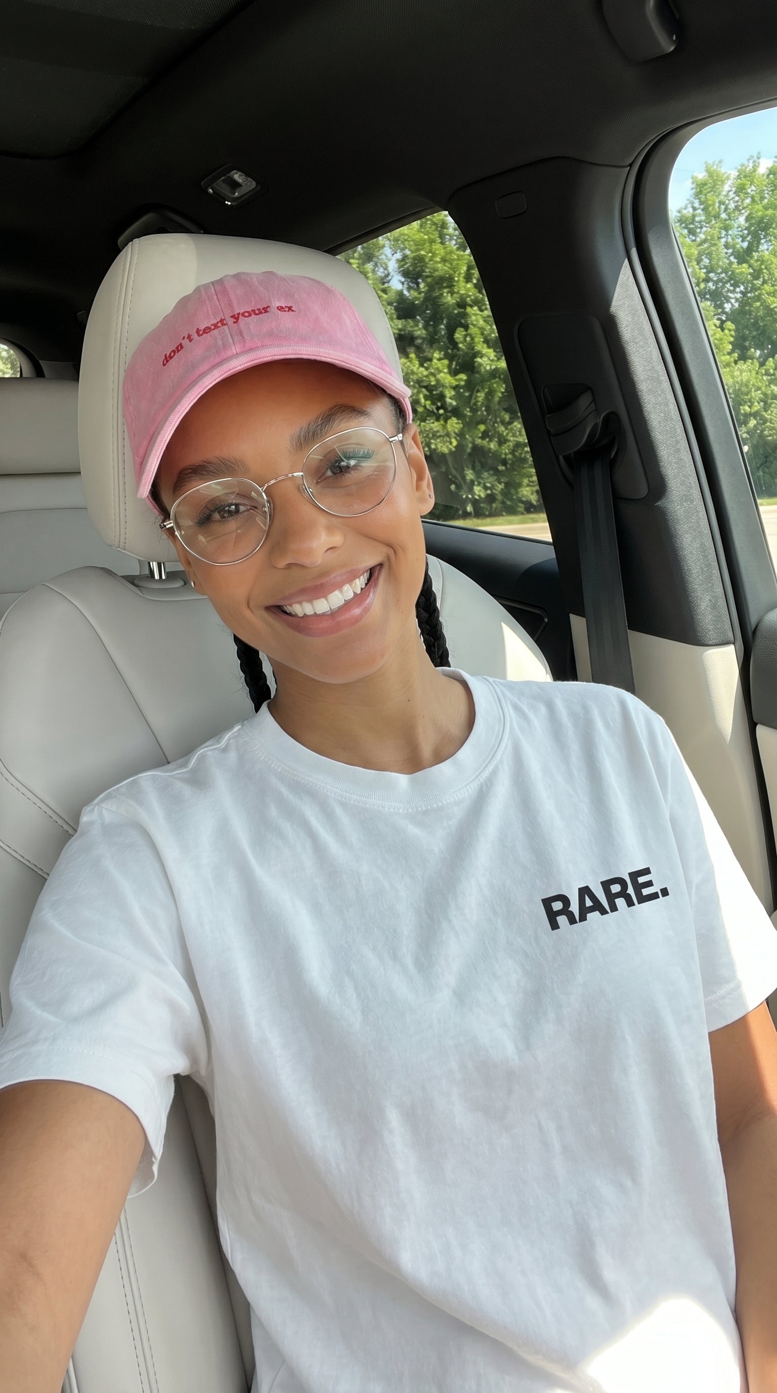 Person wearing a pink cap and white t-shirt with 'RARE' text inside a car.